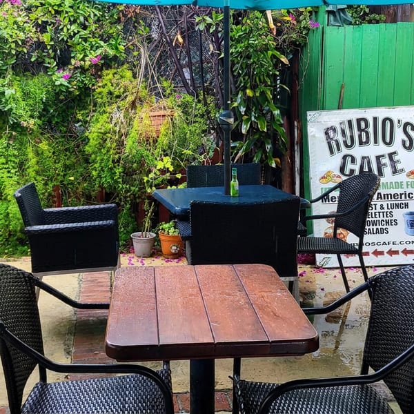 Outdoor Rubico’s Cafe patio with black wicker chairs, wooden tables, lush greenery, and a sign advertising Mediterranean and American food.