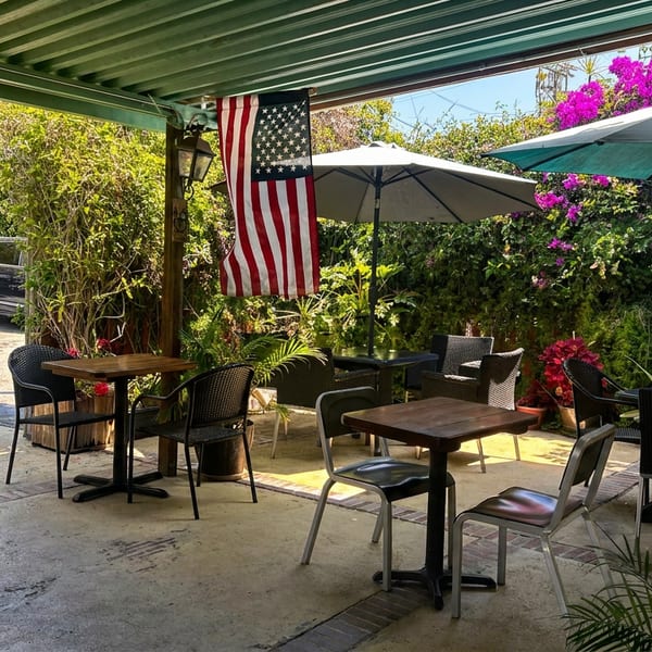 Outdoor restaurant patio with tables, chairs, umbrellas, lush flowering plants, and an American flag.