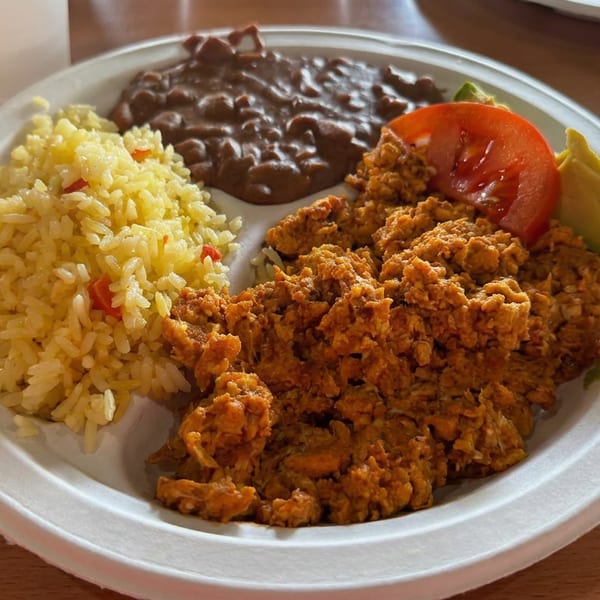 Plate of crispy fried meat with yellow rice, black beans, tomato, avocado, and a foil-wrapped item on a restaurant table.