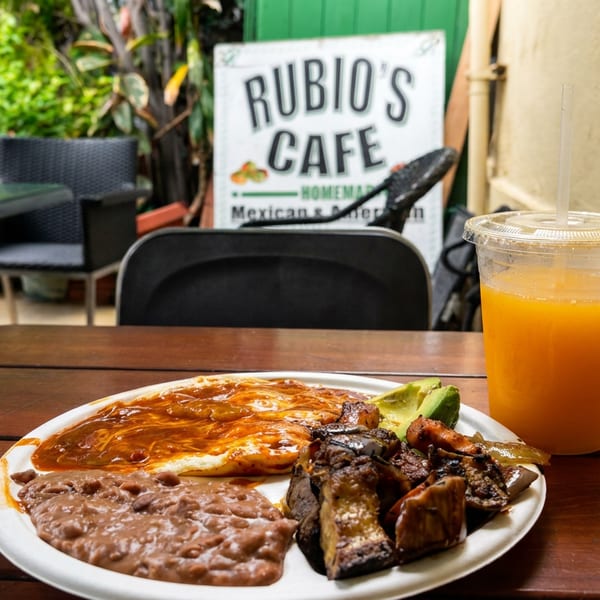 Mexican breakfast plate with eggs, beans, grilled meat, and avocado beside an orange drink at Rubio’s Café patio.