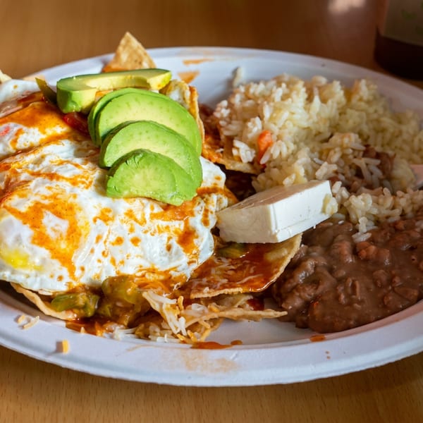Mexican-style plate with fried eggs, avocado slices, rice, refried beans, tortillas, and white cheese on a wooden table.
