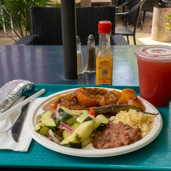 Mexican meal with rice, refried beans, grilled vegetables, cucumber salad, and a red drink on an outdoor patio table.