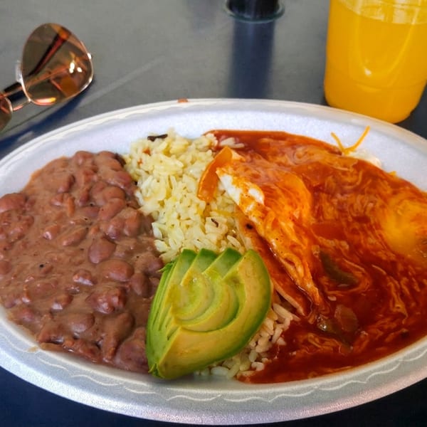 Mexican plate with enchiladas in red sauce, refried beans, rice, and avocado, alongside an orange drink and sunglasses.